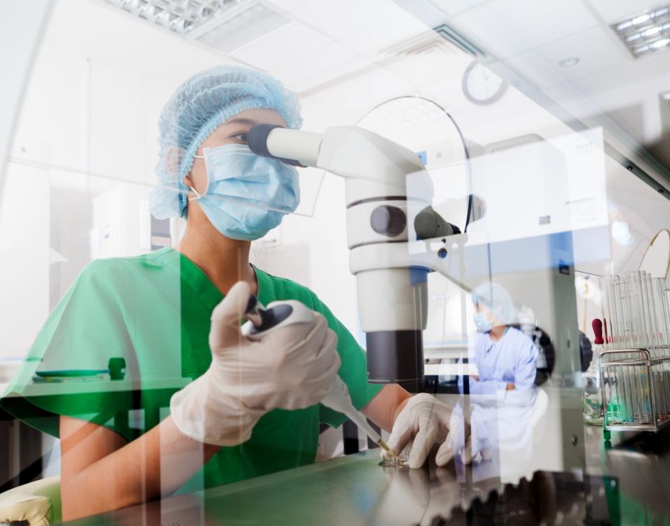 A lab researcher using a pipette to place liquid on a sample tray beneath a high-powered microscope.