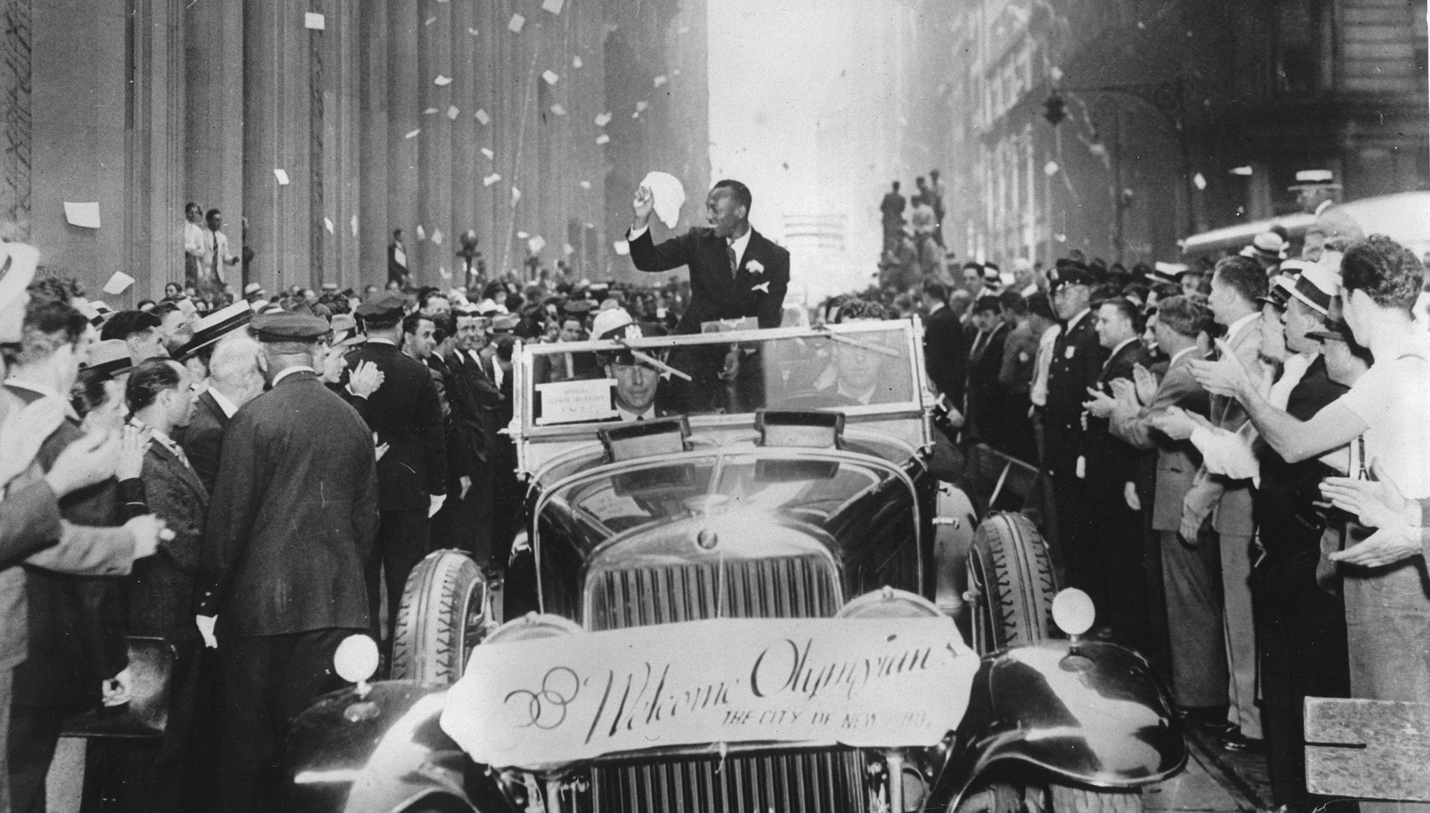 Jesse Owens driving in an open top car along Broadway in New York with thousands of people on the streets to see him