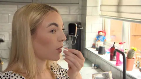 Alex Dunlop/BBC Gemma Cutler taking an over-the-counter painkiller in a kitchen