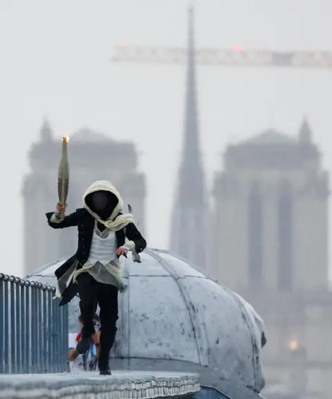 Peter Cziborra/Pool/Getty Images A masked torchbearer runs atop the Musee d'Orsay with the Notre-Dame-de-Paris cathedral in the background during the Opening Ceremony of the Olympic Games Paris 2024 on July 26, 2024