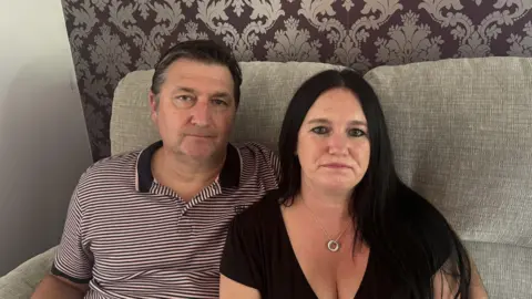 George King/BBC Nick and Vicky look directly at the camera, photographed sitting on a beige sofa. Nick is wearing a red and white striped polo neck, and Vicky is in a black T-shirt and wearing a silver necklace. 