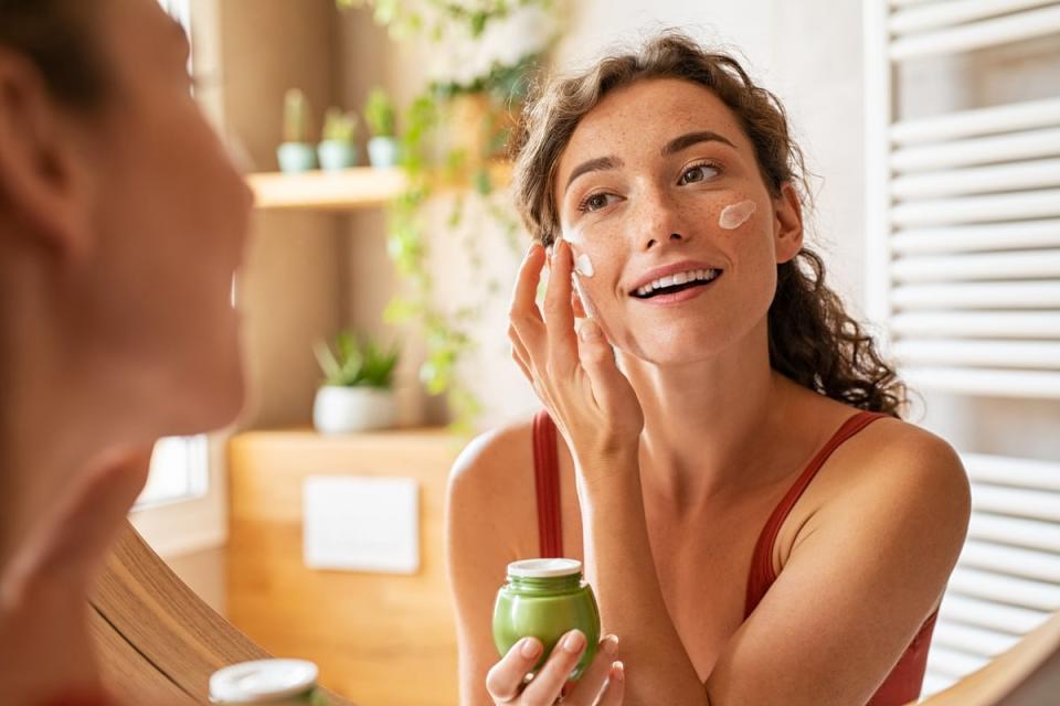 A person smiles while using a mirror to apply facial cream.