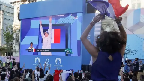 Getty Images A woman in the foreground cheers as swimmer Léon Marchand appears on a big screen