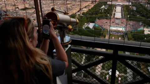 Getty Images A woman uses a telescope on the Eiffel Tower to watch a volleyball match in a stadium below