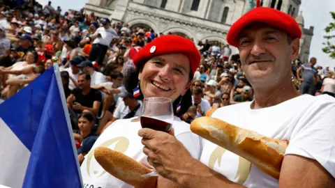 Getty Images A man and woman wearing French berets hold red wine and baguettes in front of a crowd