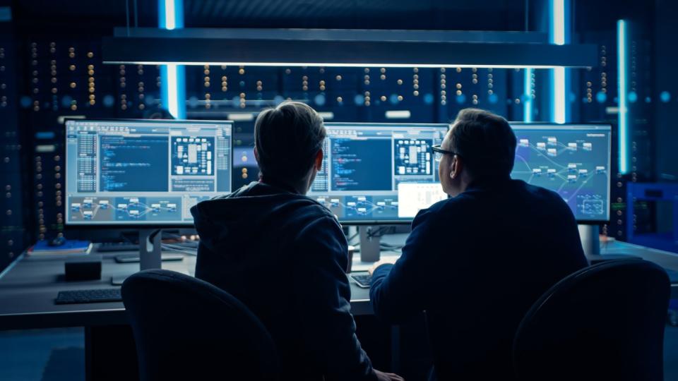 Two employees looking at copious amounts of data displayed on multiple computer screens. 