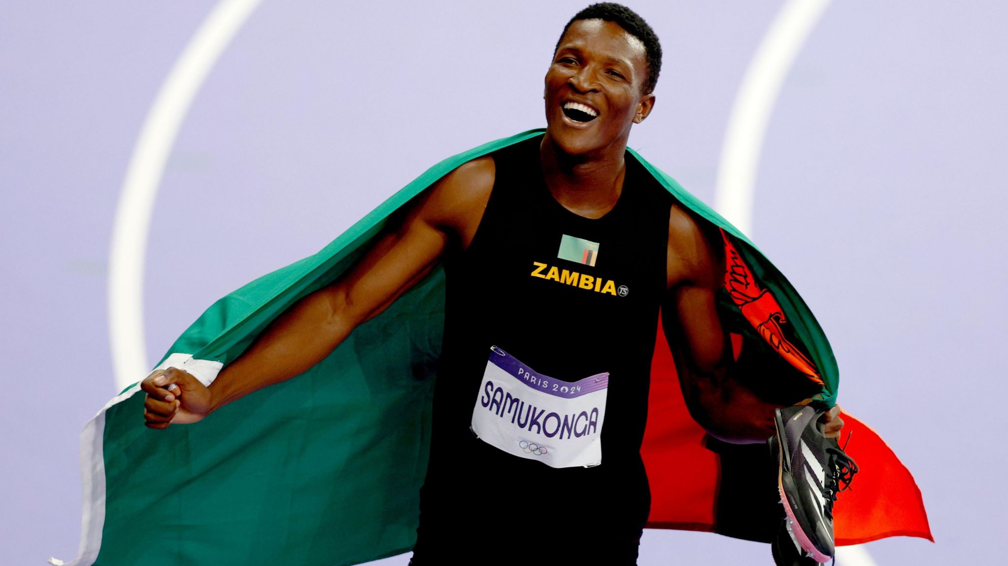 Muzala Samukonga simles as he celebrates winning a bronze medal, trailing a Zambia flag behind him while holding his spikes in his left hand