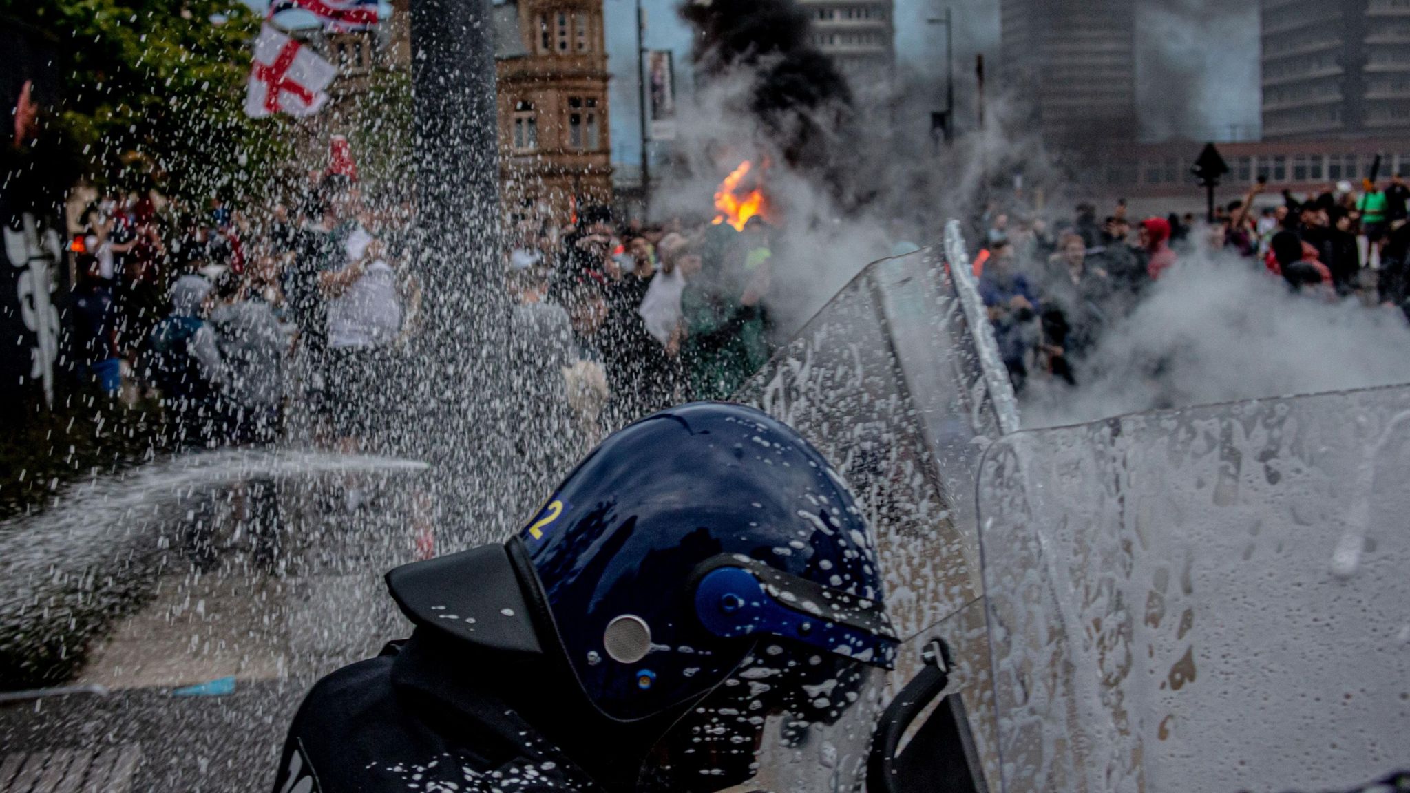 A police officer holds up a riot shield with a crowd of protestors in the background