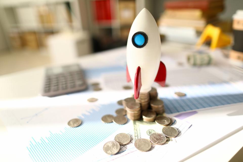 A toy rocket prepping for launch atop messy stacks of coins and paperwork displaying financial information.