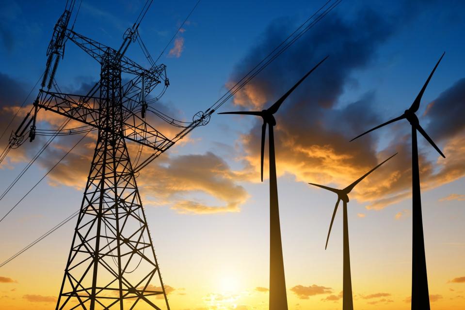 Three wind turbines next to an electrical tower, with the sun rising in the horizon. 