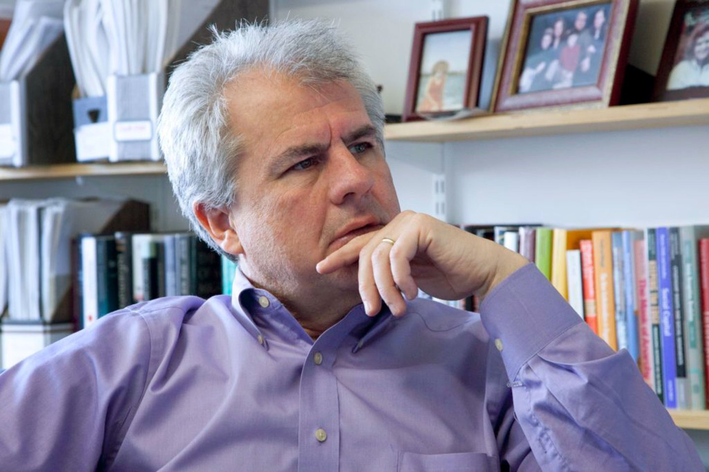 White-haired man strokes chin in front of bookshelf