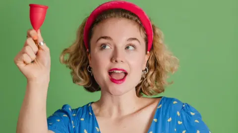 Instagram/Bella Humphries Comedian Bella Humphries wearing a red headband with mouth slightly open holding a mooncup to the left and looking up towards it. 