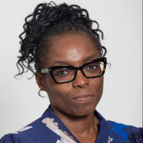 Grainge Photography Dr Lade Smith, wearing black-framed glasses and a blue top, looks directly to camera.