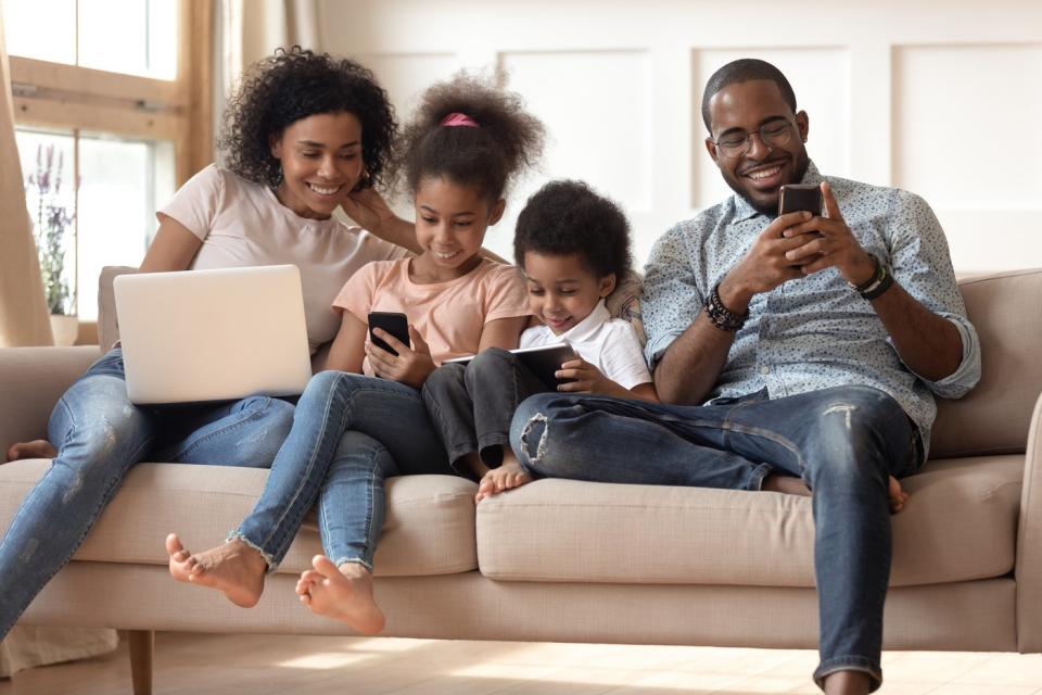 A family of four seated on a couch, each of whom are engaged with their own wireless device. 