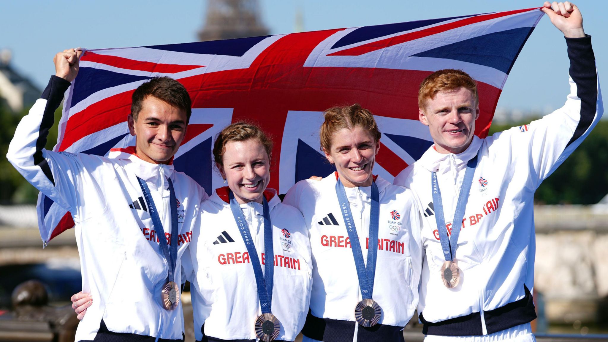 Alex Yee, Georgia Taylor-Brown, Beth Potter and Sam Dickinson pose with a Great Britain flag after winning bronze in the mixed relay triathlon at the Paris 2024 Olympics