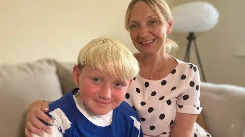 BBC Hector and Natalie sitting on a sofa at home. They both have blonde hair and are smiling. He is wearing a blue football shirt. She is wearing a polka dot top.