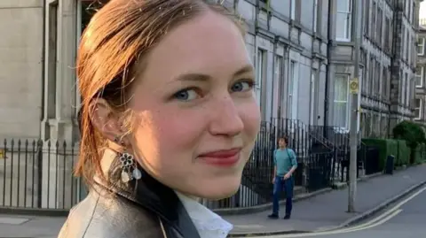 Cordelia Ostler Rhiannon has brown, tied back hair and is wearing a black leather coat and white blouse. She is turned to the camera and is smiling. Behind her are a row of houses in Edinburgh where she was studying at university.