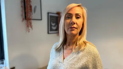 Michelle Sword looking at the camera. She is at her home. Some images and decorations could be seen on the wall behind her.