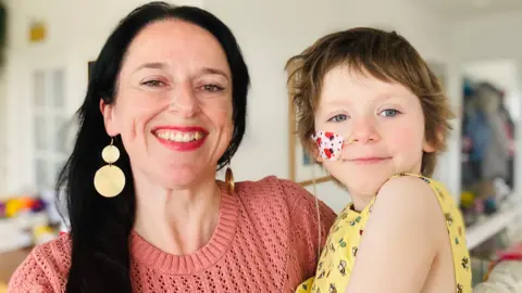 Libby Ashmore holds her daughter Annabel. Libby has long dark hair and is wearing a pink jumper. Annabel is in a yellow dress, has short dark hair and a tube coming from her nose concealed by a plaster with a ladybird pattern on it.