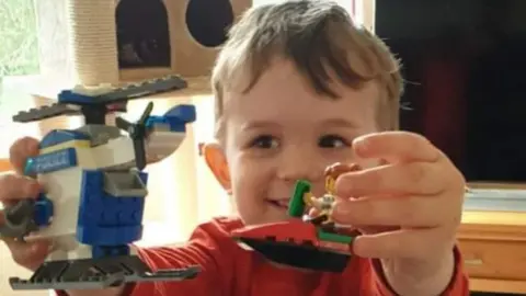 Family photo Alexander smiles at two Lego figures: one a police helicopter, the other a figure on a boat. He has brown hair and is wearing a red long sleeved top in a lounge in front of a TV and cat gym. 