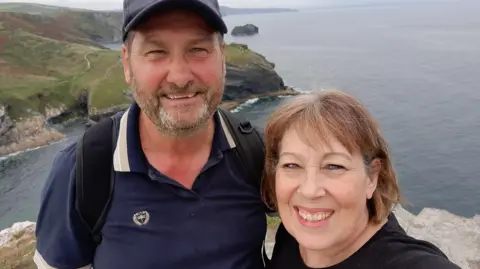 Lynda Sawyer Kim and Lynda Sawyer smile for a selfie as they stand on cliffs with a view of the sea and coastline behind them. Kim, who has a beard, is wearing a dark blue polo shirt, a dark blue baseball cap and a black backpack. Kim, whose hair is cut in a bob, is wearing a black top.