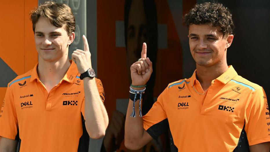 McLaren's Oscar Piastri and Lando Norris hold up their index fingers while stood next to each other and smiling