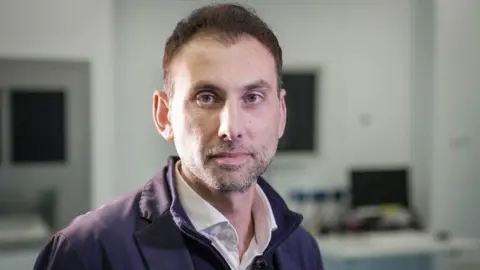 Anastasios Giamouriadis, man with black hair and grey stubble, wearing open-necked shirt and blue jacket, looking at camera, with computer equipment in background.