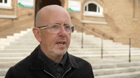 Sean O'Neill, Maeve Boothby-O'Neill's father, is standing at the bottom of stone steps talking to the camera. He is wearing black and a pair of glasses.