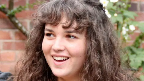 Family handout/PA Media Maeve Boothby-O'Neill is looking to the side of the camera and is smiling. She is standing outside near a tree and has long, dark, wavy hair and brown eyes.