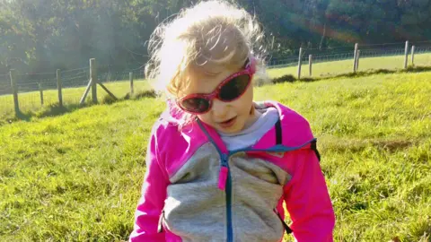 Lisa Hawker Jaydi wearing a pink and grey zip up hoodie with pink sunglasses while out on a sunny day in a grassy field with trees in the background