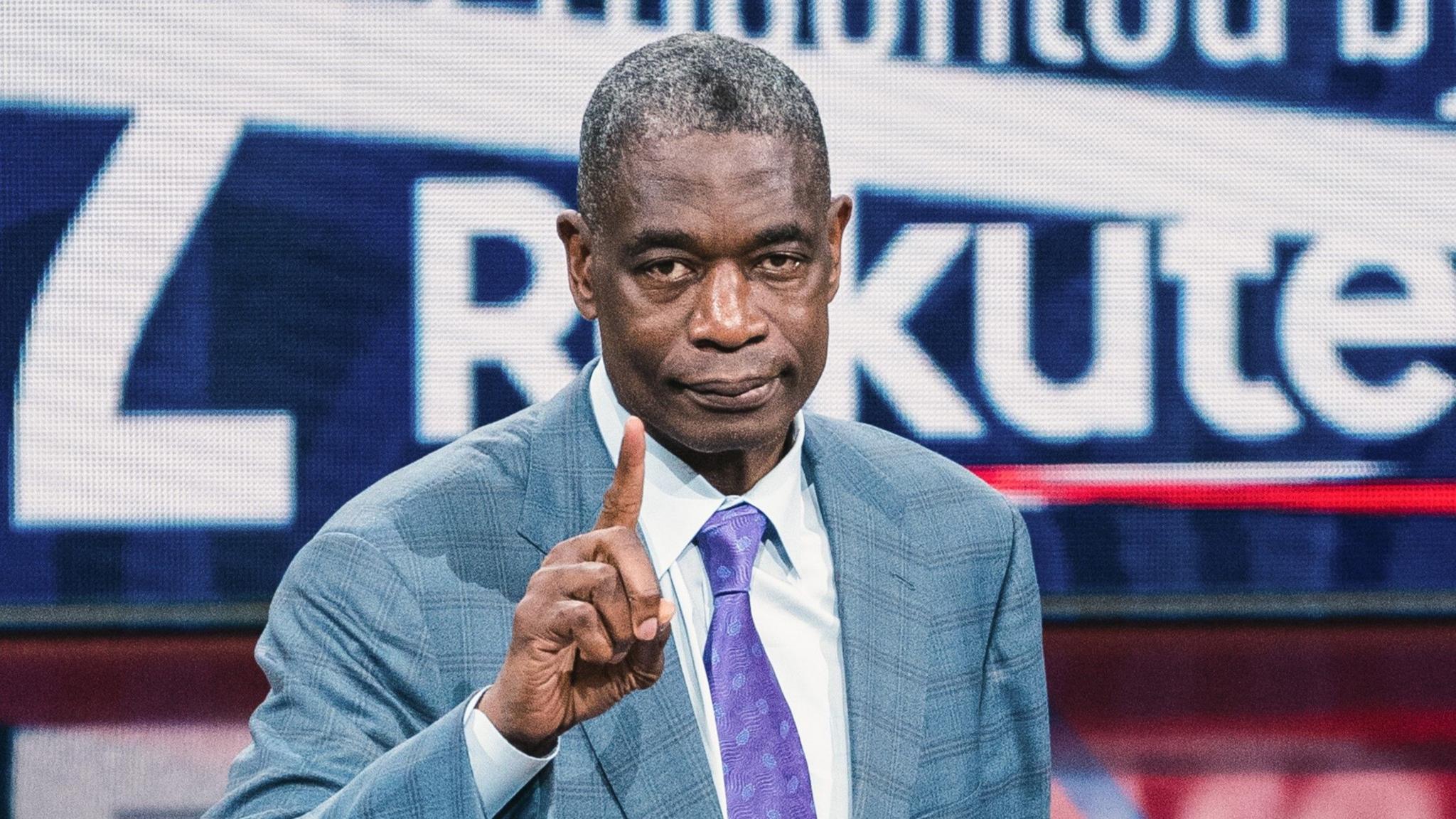 Dikembe Mutombo, wearing a light blue suit with a purple tie, raises an index finger towards the camera