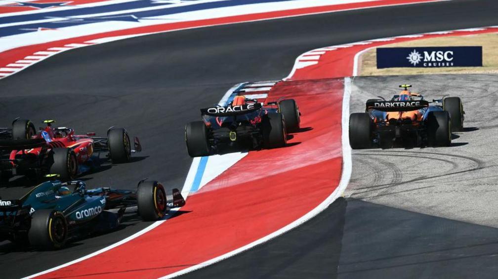 A rear view of Max Verstappen and Lando Norris leaving the track on the first lap as Charles Leclerc prepares to pass both and take the lead