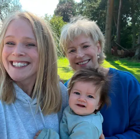 Sally Magnusson Selfie of Sally Magnusson (right) in a navy blue top next to her daughter Anna Lisa who is wearing a grey hoodie. Anna Lisa is holding her daughter Remy who has dark hair. The sun is shining on the bright green grass and trees behind them and they are all smiling at the camera.