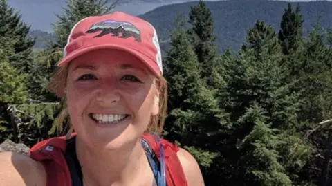 HANDOUT Ellie Lambert, pictured in a sunny forest in Canada and wearing a red cap with a bear on it and hiking clothes, smiles in a selfie 