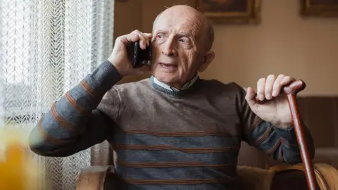 Getty Images An elderly man holds a phone to his ear as he makes a call while looking out the window at home. He's sat on a chair near the window, which is covered by light white transparent curtains, and wears a jumper over a shirt, and holds a walking stick in his free hand.