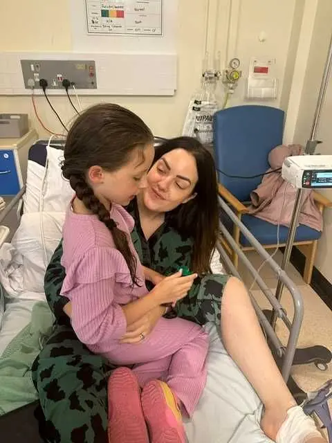 Anna Cooper Lady on a hospital bed, dressed in green patterned pyjamas, smiling at her young daughter who is sitting on her lap. 