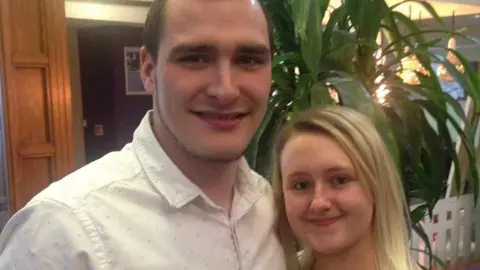 HANDOUT Smiling Chris Taylor, who has brown hair and a beard and is wearing a white shirt, is pictured with his partner Jemma, who has long blonde hair and minimal make-up, in the foyer of a venue