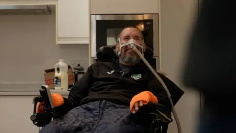 Nik smiles at his family behind the camera as he sits in a motorised wheelchair with a breathing mask across his face