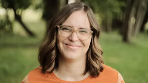 Clare Smyrell A smiling Clare Smyrell with mid-length light brown hair, glasses, wearing an orange top, with grass and trees seen blurred in the background