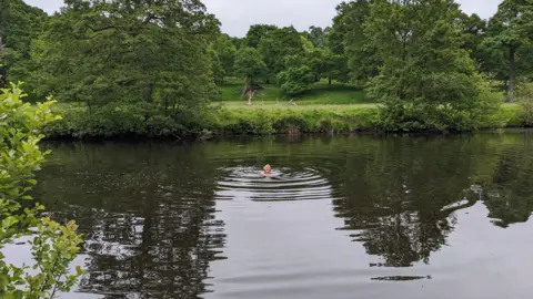 Jo Broughton A woman swimming in a river by a tow path with greenery surrounding the bank