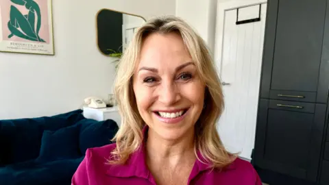 BBC A woman with blonde hair and pale pink lipstick smiles widely at the camera