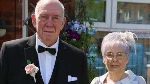 Family photograph Roy and Susan Pollit pose for a photograph together. Roy wears a formal black suit with a black bow-tie and a pink flower in his button hole. Susan wears a white fascinator and a light grey outfit.