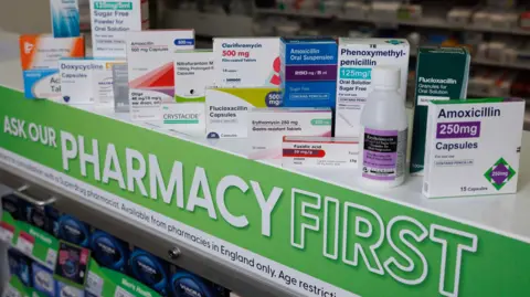 PA 24 different types of medications that Superdrug is now able to supply to patients under the Pharmacy First service. They are all lined up on a shelf with "Ask our Pharmacy First" printed in white letters on a green sign.