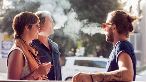 Getty Images Three people are standing together outdoors - one woman has a cigarette in her hand while the other two men are blowing tobacco smoke out of their mouths