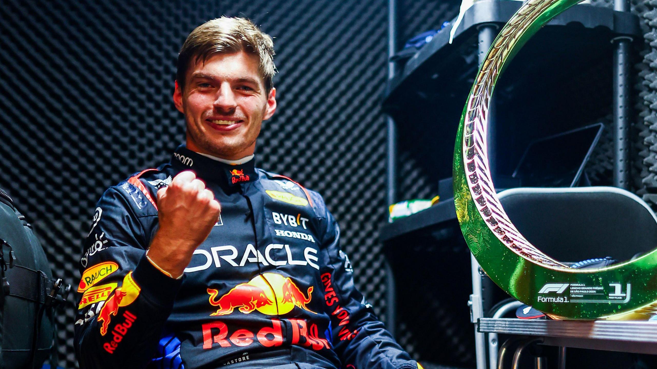 A smiling Max Verstappen is sat down as he clenches his right hand in celebration after winning the Sao Paulo Grand Prix. The winning trophy is to the right of him