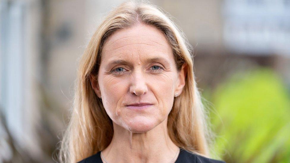 Labour MP Kim Leadbeater looks straight at the camera, in front of a blurred background. She has long hair, and is wearing a black top.