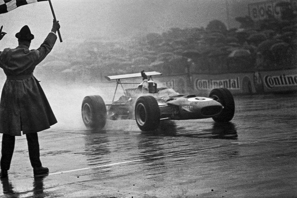 A man in a raincoat is stood on the Nurburgring circuit as he waves the chequered flag as Jackie Stewart crosses the line to win the 1968 German Grand Prix. Stewart has raised his right arm in celebration