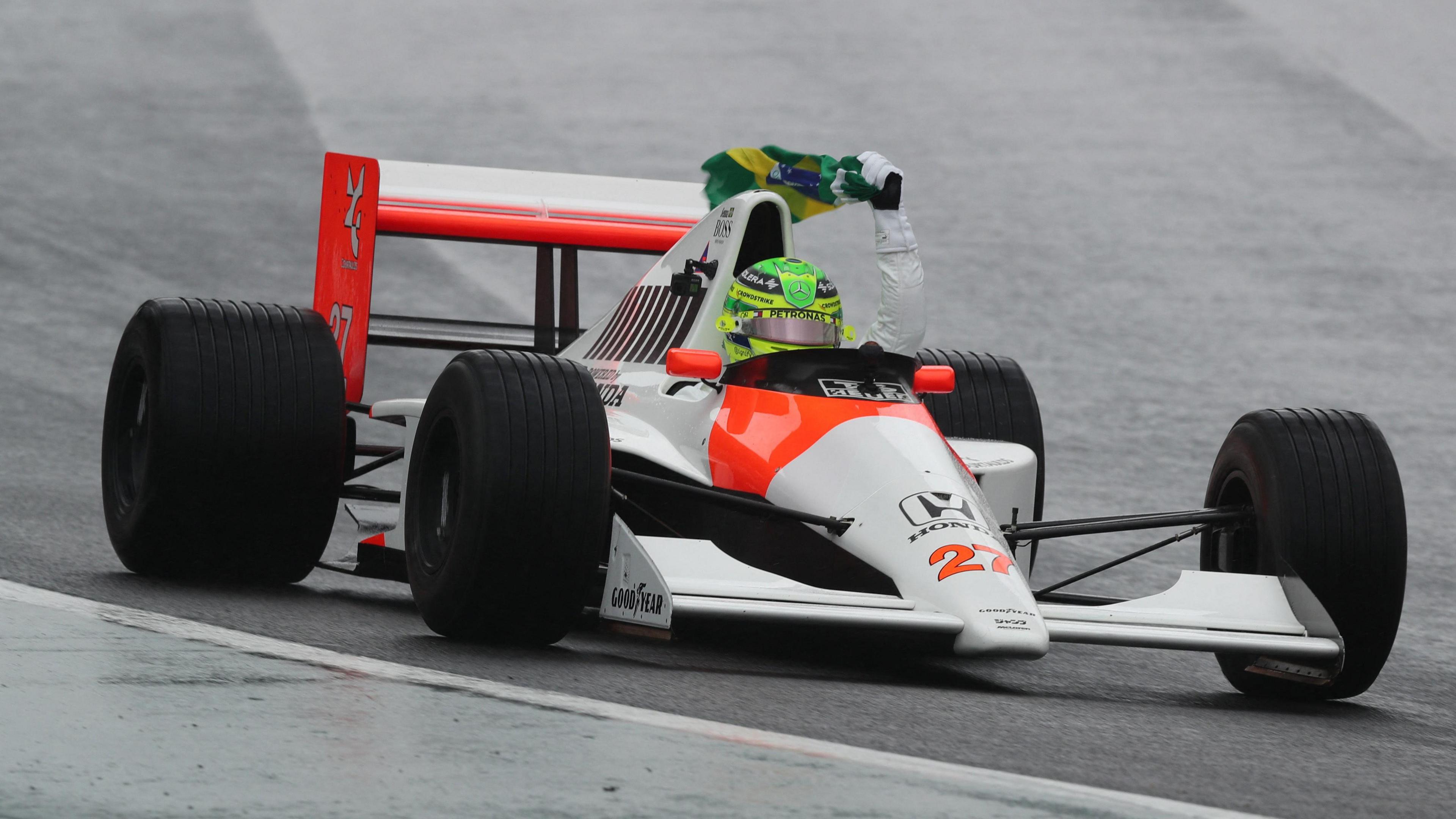 Lewis Hamilton drives Ayrton Senna's 1990 McLaren-Honda at Interlagos, holding a Brazilian flag above his head with his left hand while doing so