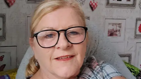 Wendy Swain Wendy Swain at home. She has glasses on and is smiling. She has blonde hair, pulled behind her head in a ponytail and has blue eyes. Her mouth is slightly open. Behind her on a wall decorated with wallpaper with grey writing, are a series of framed pictures.