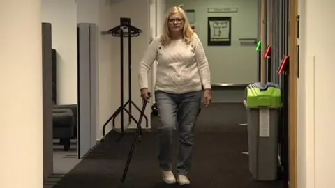 A woman in a white jumper, blue jeans and white trainers holding a walking stick while slowly walking down a corridor with a slight limp. The corridor has dark grey carpet and white walls with a black metal clothes hanger on one side and what look like grey bins with green and blue lids on the other side.